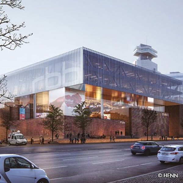 Moderne Architektur eines Gebäudes mit Glasfassade und ziegelsteinernen Elementen, umgeben von Bäumen und einem Parkplatz. Das Bild zeigt die Fassade im Vordergrund, in einer städtischen Umgebung, bei Tageslicht.