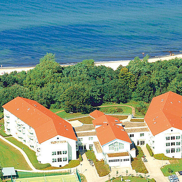 Luftaufnahme eines Gebäudekomplexes mit roten Dächern, umgeben von Bäumen, nahe einem Wasserlauf mit Sandstrand. Im Hintergrund sind Menschen am Strand zu sehen. Die Anlage liegt in einer grünen Umgebung und hat verschiedene Gebäude.