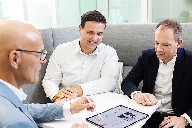 Drei Männer sitzen an einem Tisch und betrachten gemeinsam ein Tablet. Der Mann links trägt eine Brille und einen Anzug, während die anderen beiden in lässigerer Kleidung sind. Sie scheinen über den Inhalt des Tablets zu diskutieren. Der Hintergrund ist modern und hell.