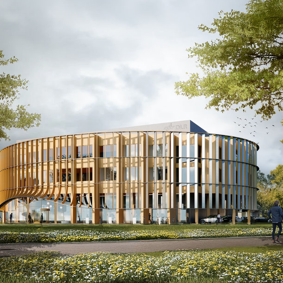 Ein modernes, rundes Gebäude mit Holzfassade und großen Fenstern, umgeben von Grünflächen und Bäumen. Im Vordergrund gehen zwei Personen auf einem Weg. Der Himmel ist bewölkt. Blumen blühen entlang des Weges.