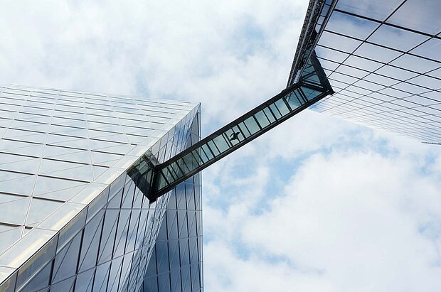 Zwei moderne Hochhäuser mit Glasfassaden, die durch einen transparenten Glaskorridor verbunden sind. Der Himmel ist bewölkt, was dem Bild eine dynamische Atmosphäre verleiht.