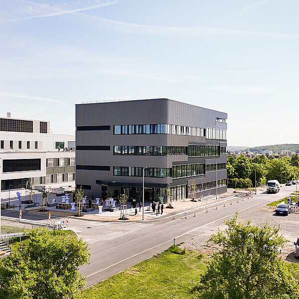 Moderne Bürogebäude mit mehreren Etagen, umgeben von grünen Flächen und einem Parkplatz. Im Vordergrund sind Autos und Fußgänger auf der Straße zu sehen. Der Himmel ist klar und blau, was auf schönes Wetter hinweist.