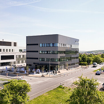 Moderne Bürogebäude mit mehreren Etagen, umgeben von grünen Flächen und einem Parkplatz. Im Vordergrund sind Autos und Fußgänger auf der Straße zu sehen. Der Himmel ist klar und blau, was auf schönes Wetter hinweist.