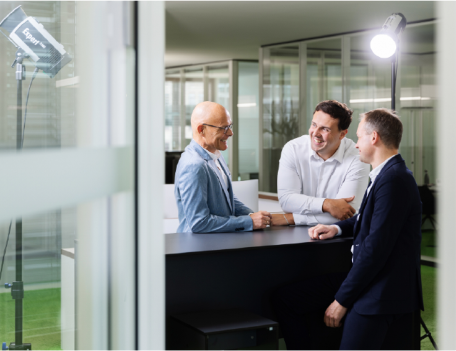 Drei Männer unterhalten sich an einem Stehtisch in einem modernen Büro. Zwei von ihnen tragen Anzüge, während der Dritte eine Brille und eine hellblaue Jacke trägt. Im Hintergrund sind Bürolandschaften und eine Lampe sichtbar.