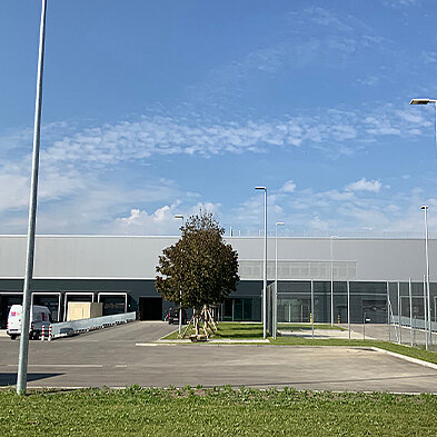 Das Bild zeigt ein modernes Gebäude mit einer grauen Fassade und großen Fenstern. Vor dem Gebäude befindet sich eine gepflasterte Fläche mit einem grünen Rasenstreifen. Ein Baum steht in der Nähe, und es sind einige parkende Lieferwagen sichtbar. Der Himmel ist klar und blau.