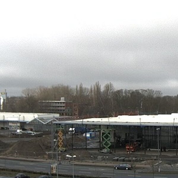 Blick auf eine Baustelle mit mehreren Baugeräten und Maschinen. Im Hintergrund sind Bäume und ein mehrstöckiges Gebäude zu sehen. Der Himmel ist bewölkt und die Umgebung wirkt städtisch. Einige Autos fahren auf der Straße entlang.