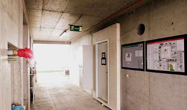 Innenansicht eines langen, schlichten Flurs mit Betonwänden. An der linken Seite ist ein rotes Rohr sichtbar, während auf der rechten Seite ein Informationsboard und eine geschlossene Tür zu sehen sind. Am Ende des Flurs führt Licht nach draußen. Ein Notausgangsschild hängt an der Decke.