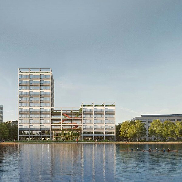 Blick auf moderne Gebäude am Ufer eines Gewässers, umgeben von Bäumen. Die Architektur ist vielfältig und zeitgenössisch, mit einer Mischung aus niedrigen und höheren Bauwerken. Der Himmel ist klar und reflektiert sich auf der Wasseroberfläche.