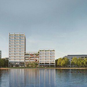 Blick auf moderne Gebäude am Ufer eines Gewässers, umgeben von Bäumen. Die Architektur ist vielfältig und zeitgenössisch, mit einer Mischung aus niedrigen und höheren Bauwerken. Der Himmel ist klar und reflektiert sich auf der Wasseroberfläche.