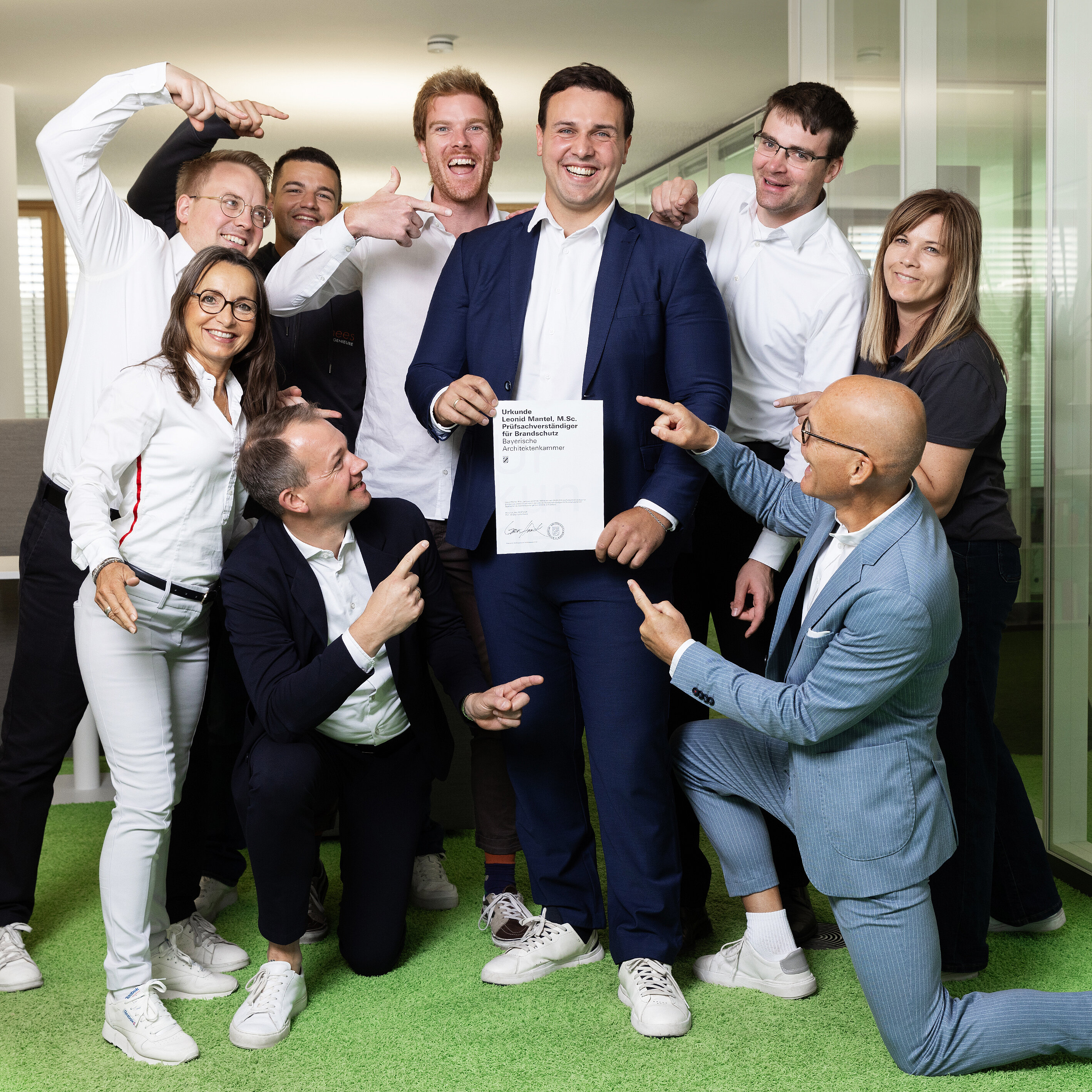 Eine Gruppe von sieben Personen steht und kniet um einen Mann in Anzug, der stolz ein Dokument hält. Alle zeigen auf ihn und lächeln, während sie in einem modernen Büro mit grünem Teppich stehen.