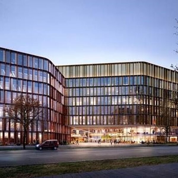 Moderne, mehrstöckige Gebäude mit großen Glasflächen und einer geschwungenen Fassade. Die Architektur kombiniert Holz und Glas, um ein einladendes Ambiente zu schaffen. Im Vordergrund sind Bäume und eine Straße mit Autos zu sehen. Der Himmel ist in sanften Farben des Sonnenuntergangs.