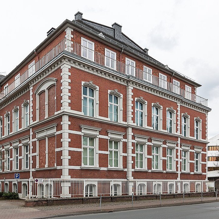 Ein mehrstöckiges, rotes Backsteingebäude mit weißen Fensterrahmen und Balkonen. Es hat eine symmetrische Fassade und bietet einen Parkplatz auf der linken Seite. Bäume im Vordergrund und eine städtische Umgebung sind ebenfalls sichtbar. Die Wolken am Himmel deuten auf eher trübes Wetter hin.