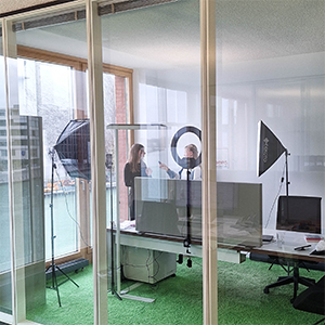 Im Bild ist ein modernes Büro mit Glaswänden zu sehen. Zwei Personen stehen in dem Raum, umgeben von Fotoapparaten und Beleuchtung. Der Boden ist mit grünem Teppich bedeckt, und große Fenster bieten einen Blick nach außen.
