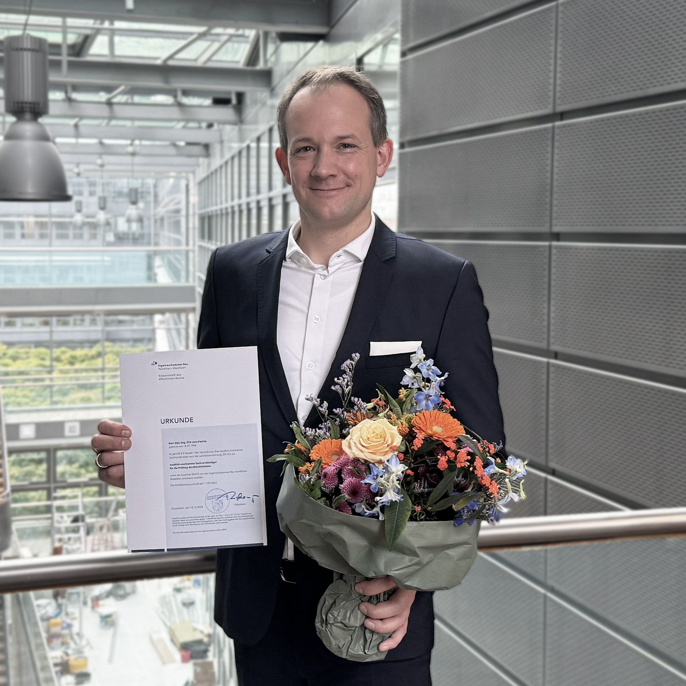 Ein Mann in einem Anzug steht in einem modernen Gebäude. In der linken Hand hält er ein Dokument mit der Aufschrift „Urkunde“, in der rechten Hand einen Blumenstrauß mit bunten Blumen. Im Hintergrund sind große Fenster und eine grüne Landschaft sichtbar.